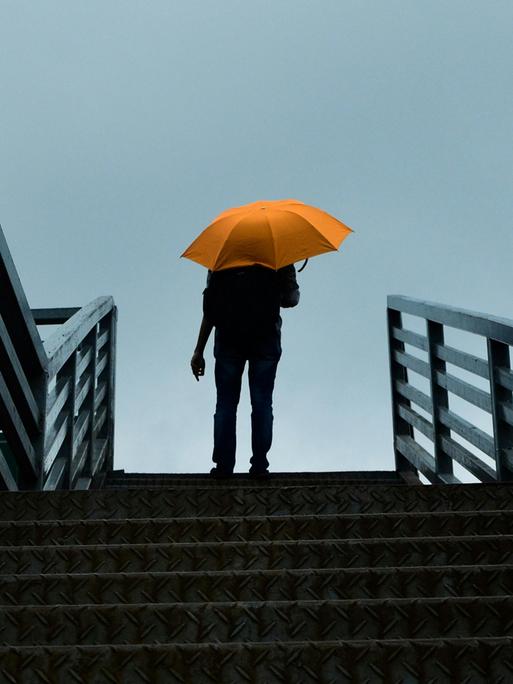 Blick auf eine große Treppe, auf der ganz oben ein Mann angelangt ist, der einen knallgelben Schirm hat, der sich leuchtend gegen den grauen Himmel abhebt.