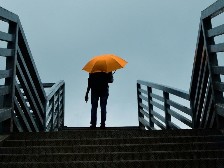 Blick auf eine große Treppe, auf der ganz oben ein Mann angelangt ist, der einen knallgelben Schirm hat, der sich leuchtend gegen den grauen Himmel abhebt.