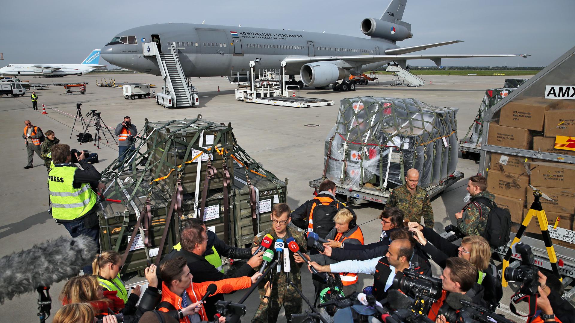 Auf einem Flughafen werden Waffen in ein Transportflugzeug geladen. Im Vordergrund richten zahlreiche Journalistinnen und Journalisten ihre Mikrofone auf einen Sprecher der Bundeswehr.