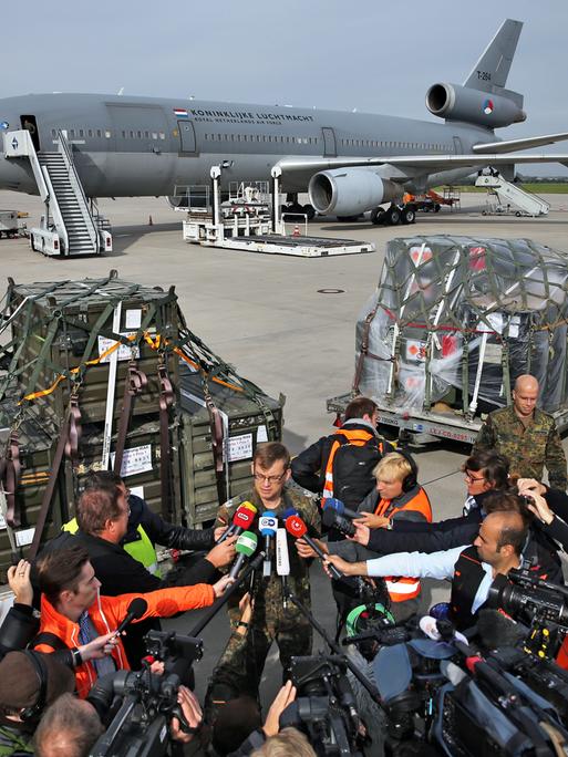 Auf einem Flughafen werden Waffen in ein Transportflugzeug geladen. Im Vordergrund richten zahlreiche Journalistinnen und Journalisten ihre Mikrofone auf einen Sprecher der Bundeswehr.