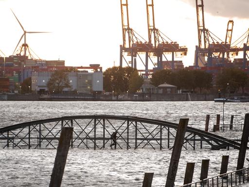 Eine Frau geht am Fischmarkt an der Elbe im Hafen während einer Sturmflut über eine unter Wasser stehende Brücke. Sturmtief „Joshua“ zog im Oktober 2025 als Herbststurm über Deutschland hinweg.