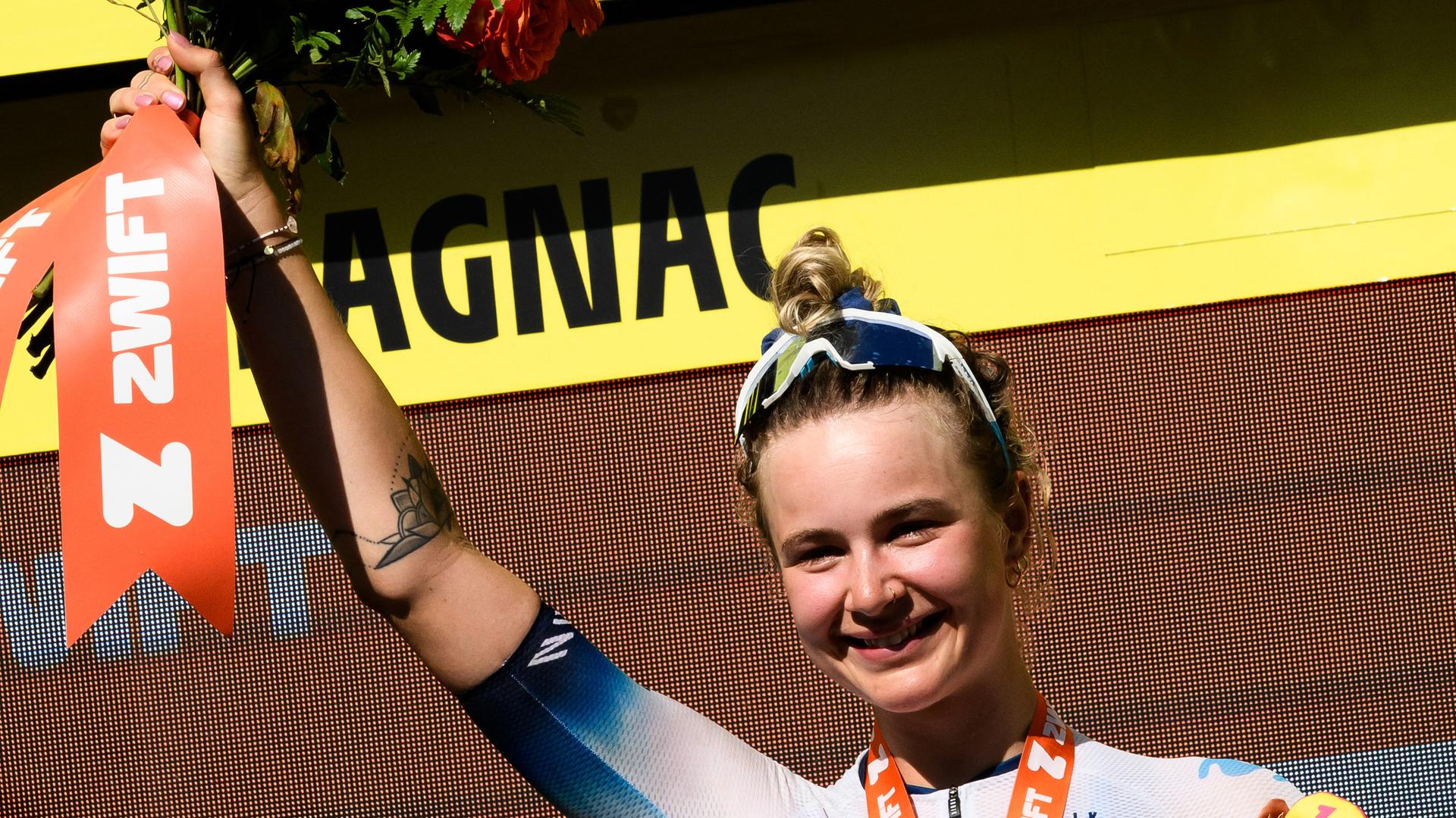 Die dänische Radfahrerin Emma Norsgaard Bjerg steht lächelnd auf dem Podium und hebt die rechte Hand in die Höhe.