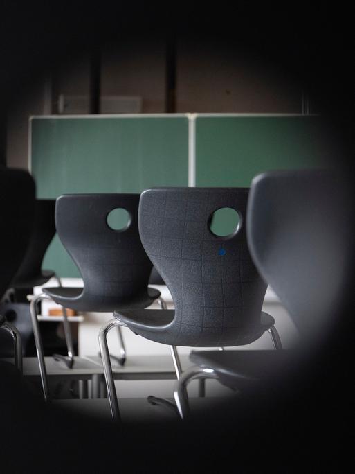 Blick durchs Schlüsseloch in ein Klassenzimmer. Zu sehen sind Stühle und eine Tafel.