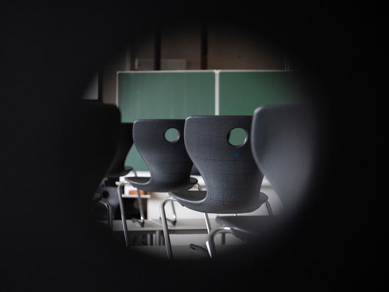 Blick durchs Schlüsseloch in ein Klassenzimmer. Zu sehen sind Stühle und eine Tafel.