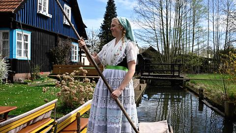 Eine blonde Frau im sorbischen Trachtenkleid steht auf einem Touristenkahn. Im Hintergrund sind Holzhäuser und eine Brücke zu sehen.