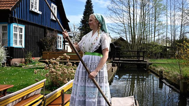 Eine blonde Frau im sorbischen Trachtenkleid steht auf einem Touristenkahn. Im Hintergrund sind Holzhäuser und eine Brücke zu sehen.
