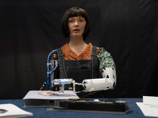 Roboter-Künstlerin Ai-Da malt Bilder mithilfe von KI in der British Library in London. Wir sehen einen Roboter mit weiblich wirkendem Gesicht. Vor ihr liegt ein Bild, auf dem sie mit dem linken Roboterarm malt. 