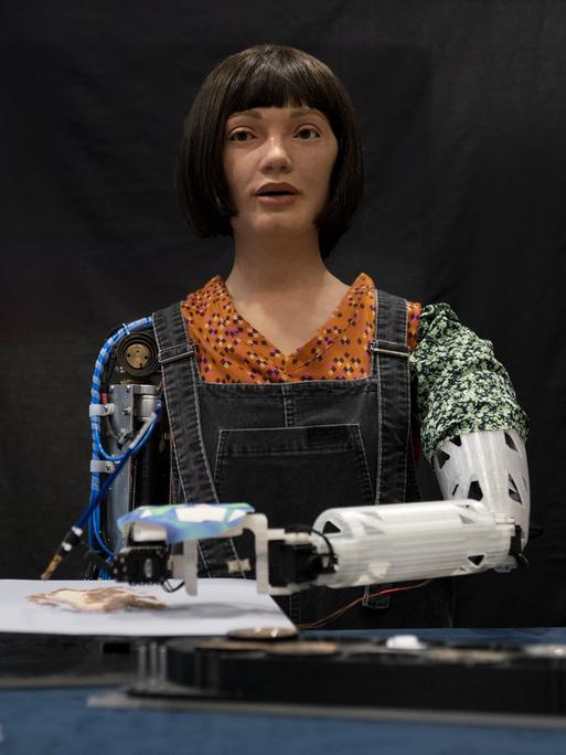 Roboter-Künstlerin Ai-Da malt Bilder mithilfe von KI in der British Library in London. Wir sehen einen Roboter mit weiblich wirkendem Gesicht. Vor ihr liegt ein Bild, auf dem sie mit dem linken Roboterarm malt. 
