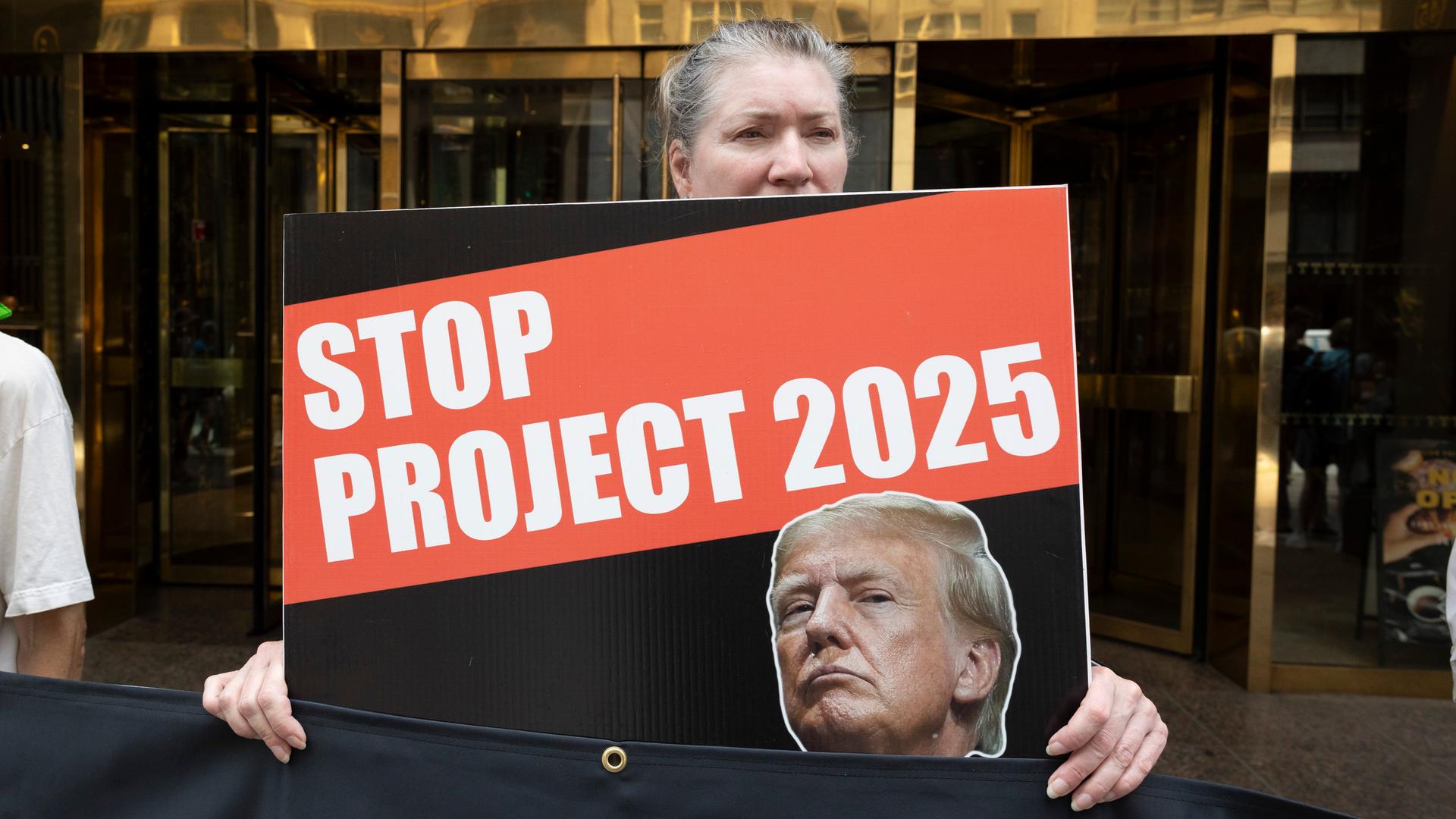 Eine grauhaarige Frau hält ein Schild mit dem Bild Trumps und der Aufschrift "Stop Project 2025". Hinter ihr der Eingang zum Gebäude mit der Aufschrift "Trump Tower".