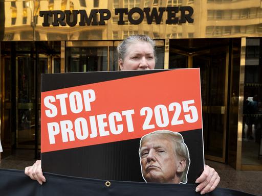 Eine grauhaarige Frau hält ein Schild mit dem Bild Trumps und der Aufschrift "Stop Project 2025". Hinter ihr der Eingang zum Gebäude mit der Aufschrift "Trump Tower".