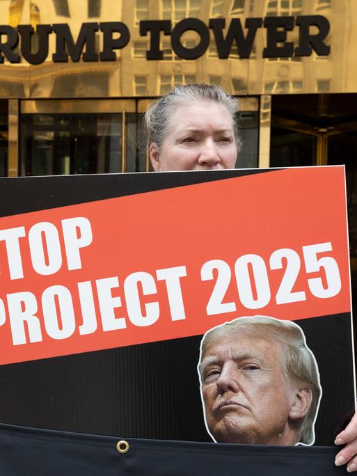 Eine grauhaarige Frau hält ein Schild mit dem Bild Trumps und der Aufschrift "Stop Project 2025". Hinter ihr der Eingang zum Gebäude mit der Aufschrift "Trump Tower".