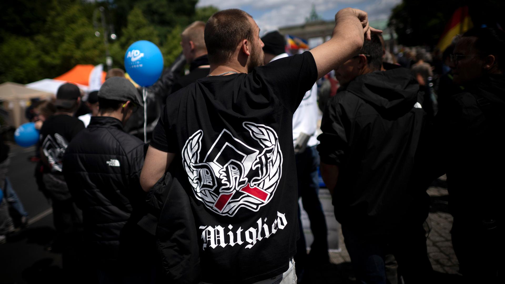 Blick auf eine Demonstration der als rechtsextrem eingestuften Partei AfD, der Querdenker-Bewegung und anderen rechten Gruppierungen. Im Vordergrund sieht man einen Demonstrant von hinten mit einem schwarzen T-Shirt mit einem Runenzeichen. 