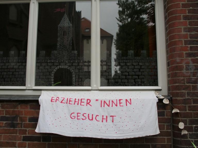 Ein Transparent auf dem geschrieben steht, "Erzieher*innen gesucht" an einer Häuserwand aus Backstein in Berlin.