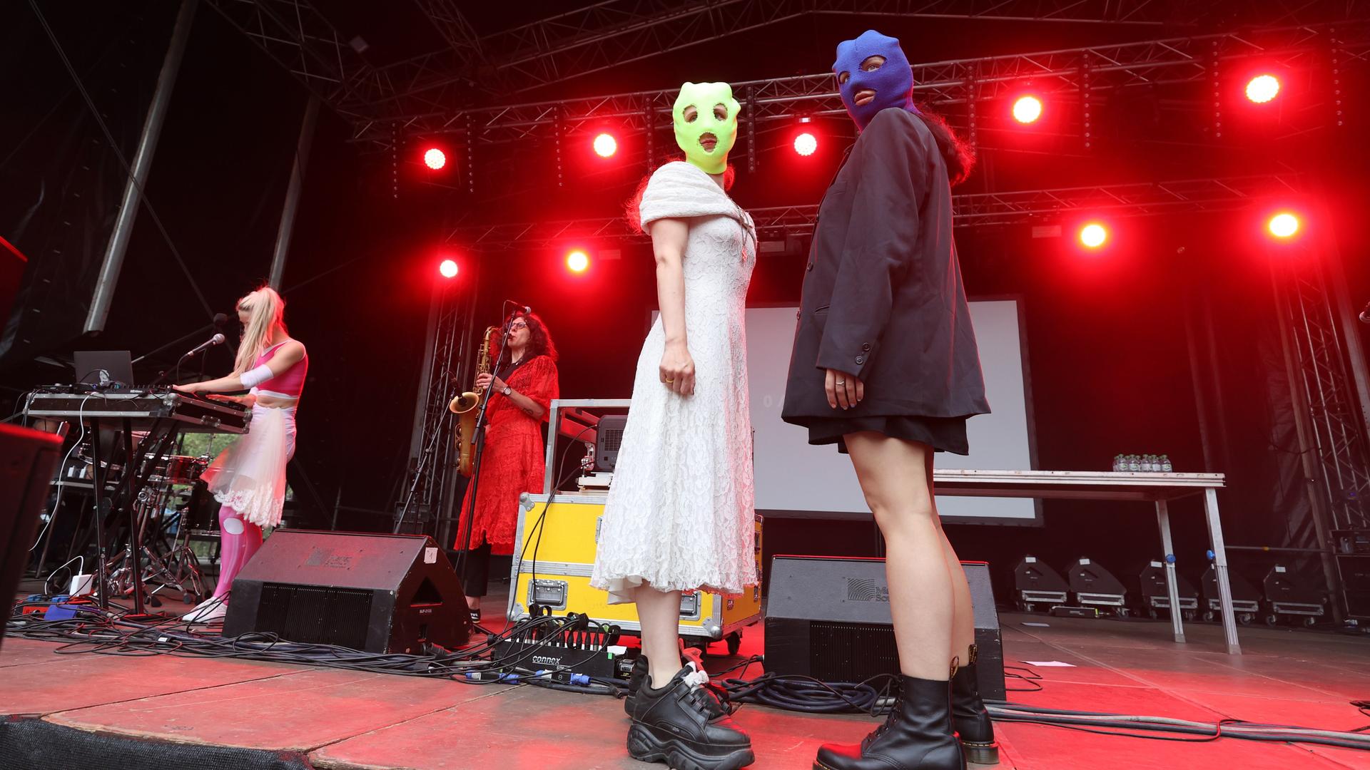 Zwei maskierte Frauen von der Band Pussy Riot stehen auf einer Bühne. Im Hintergrund eine Saxofonistin und eine Keyboarderin.