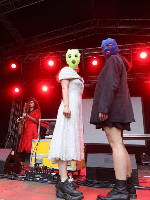 Zwei maskierte Frauen von der Band Pussy Riot stehen auf einer Bühne. Im Hintergrund eine Saxofonistin und eine Keyboarderin.