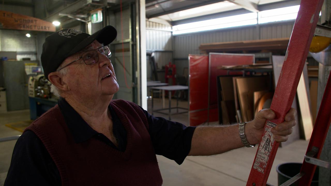 Ein älterer Mann mit Käppi und Brille steht in einer Werkstatt mit einer Holzlatte in der Hand.