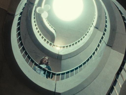 Eine Frau steht in einem spiralförmigen Treppenhaus. Der Blick geht nach oben, dort ist ein Lichtkegel zu sehen. Die Farben sind entsättigt. Die Szene hat etwas Klaustrophobisches. 