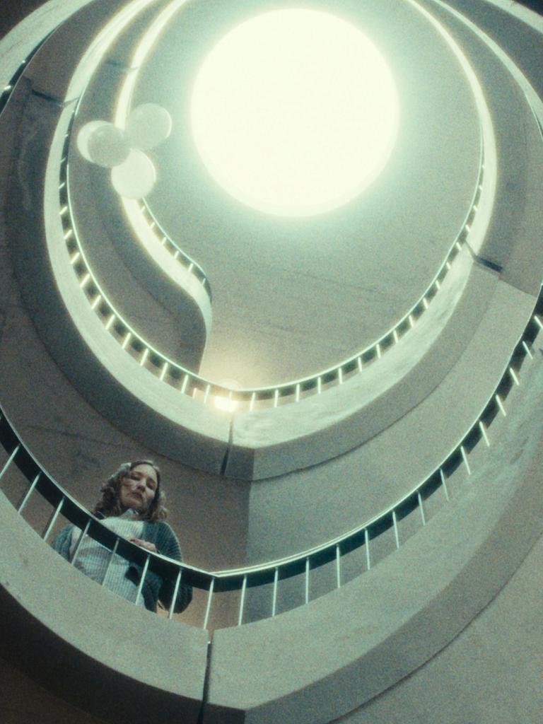 Eine Frau steht in einem spiralförmigen Treppenhaus. Der Blick geht nach oben, dort ist ein Lichtkegel zu sehen. Die Farben sind entsättigt. Die Szene hat etwas Klaustrophobisches. 