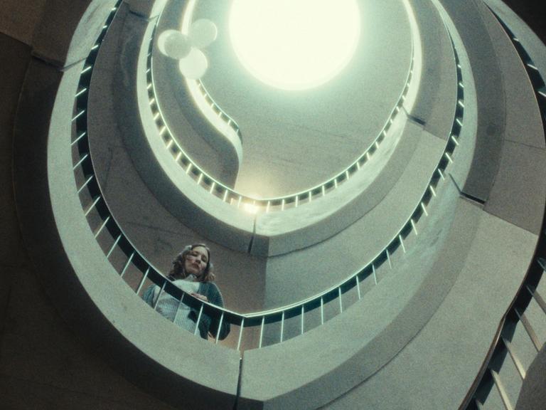 Eine Frau steht in einem spiralförmigen Treppenhaus. Der Blick geht nach oben, dort ist ein Lichtkegel zu sehen. Die Farben sind entsättigt. Die Szene hat etwas Klaustrophobisches. 