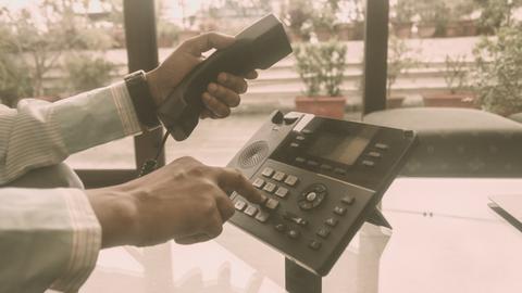 Close Up der Hände eines Mannes im Büro, der einen Telefonhörer in der einen Hand hat und mit der anderen auf dem Telefon eine Nummer wählt.