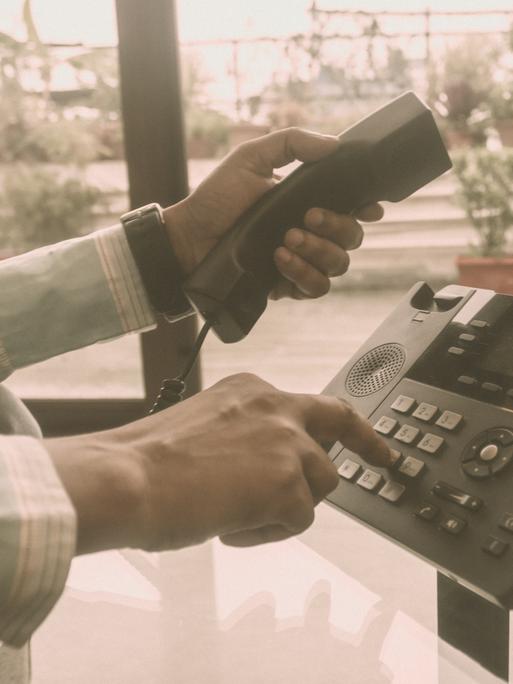 Close Up der Hände eines Mannes im Büro, der einen Telefonhörer in der einen Hand hat und mit der anderen auf dem Telefon eine Nummer wählt.