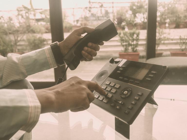 Close Up der Hände eines Mannes im Büro, der einen Telefonhörer in der einen Hand hat und mit der anderen auf dem Telefon eine Nummer wählt.