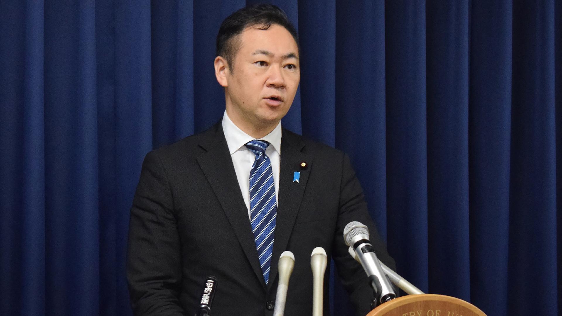Japans Justizminister Suzuki steht hinter einem Rednerpult mit mehreren Mikrofonen.