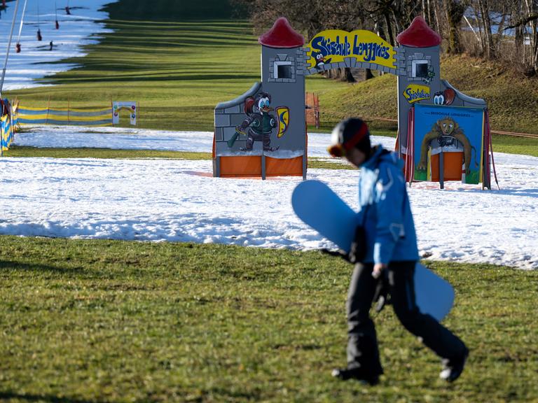 Eine Junge geht mit seinem Snowboard am Draxlhang im Skigebiet Brauneck über eine grüne Wiese. Viele Skigebiete in Bayern leiden aktuell unter Schneemangel.