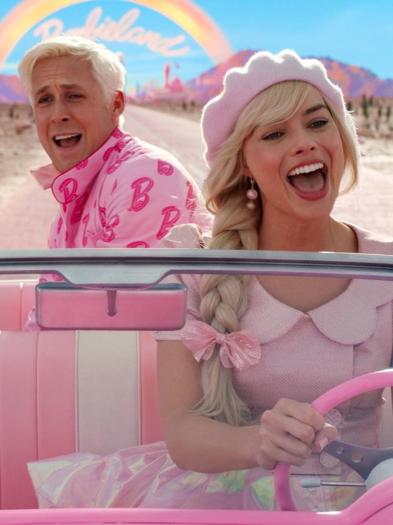 Ryan Gosling als Ken und Margot Robbie als Barbie am Lenkrad fahren grinsend in einem pinken Cabriolet durch eine wüstenartige Landschaft.