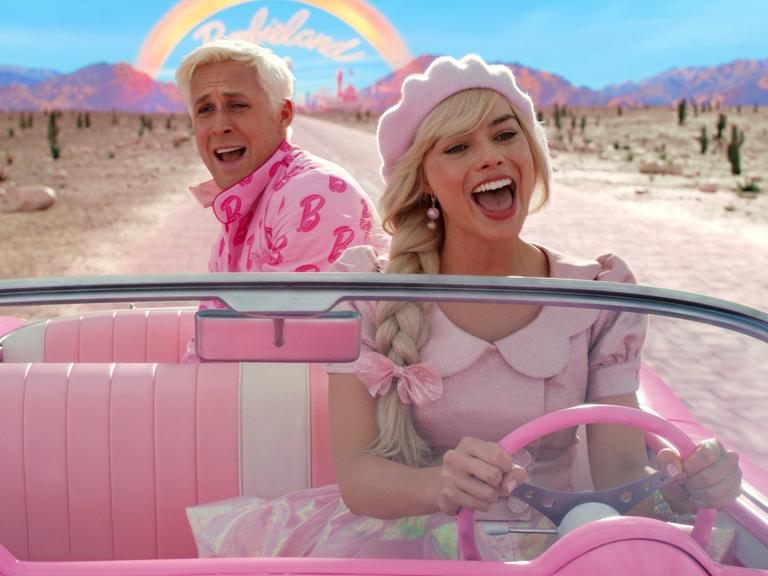 Ryan Gosling als Ken und Margot Robbie als Barbie am Lenkrad fahren grinsend in einem pinken Cabriolet durch eine wüstenartige Landschaft.