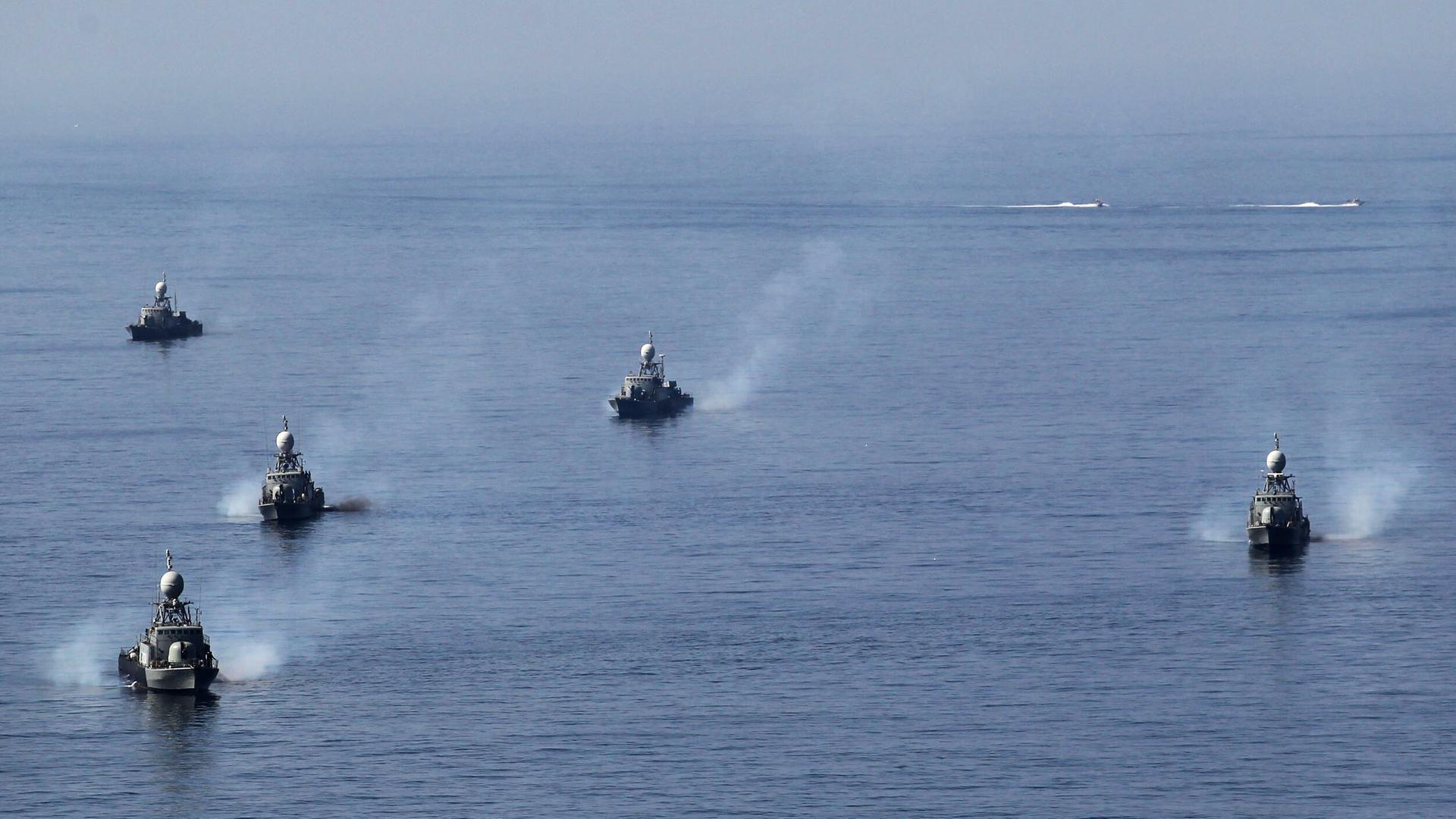 Blick aus großer Ferne: Fünf Schiffe fahren durch das Meer und ziehen Rauchschwaden hinter sich her. Man sieht sie von vorne.