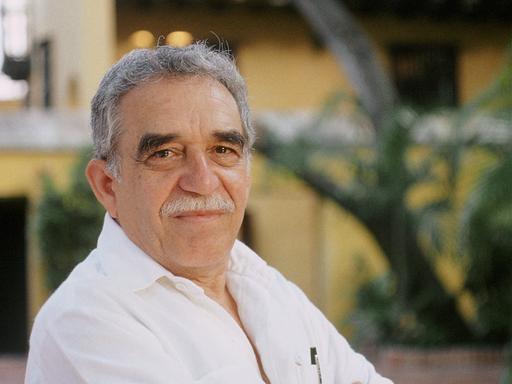 Portrait von Gabriel Garcia Marquez der vor einem gelben Gebäude in einem Innenhof mit Bäumen steht.