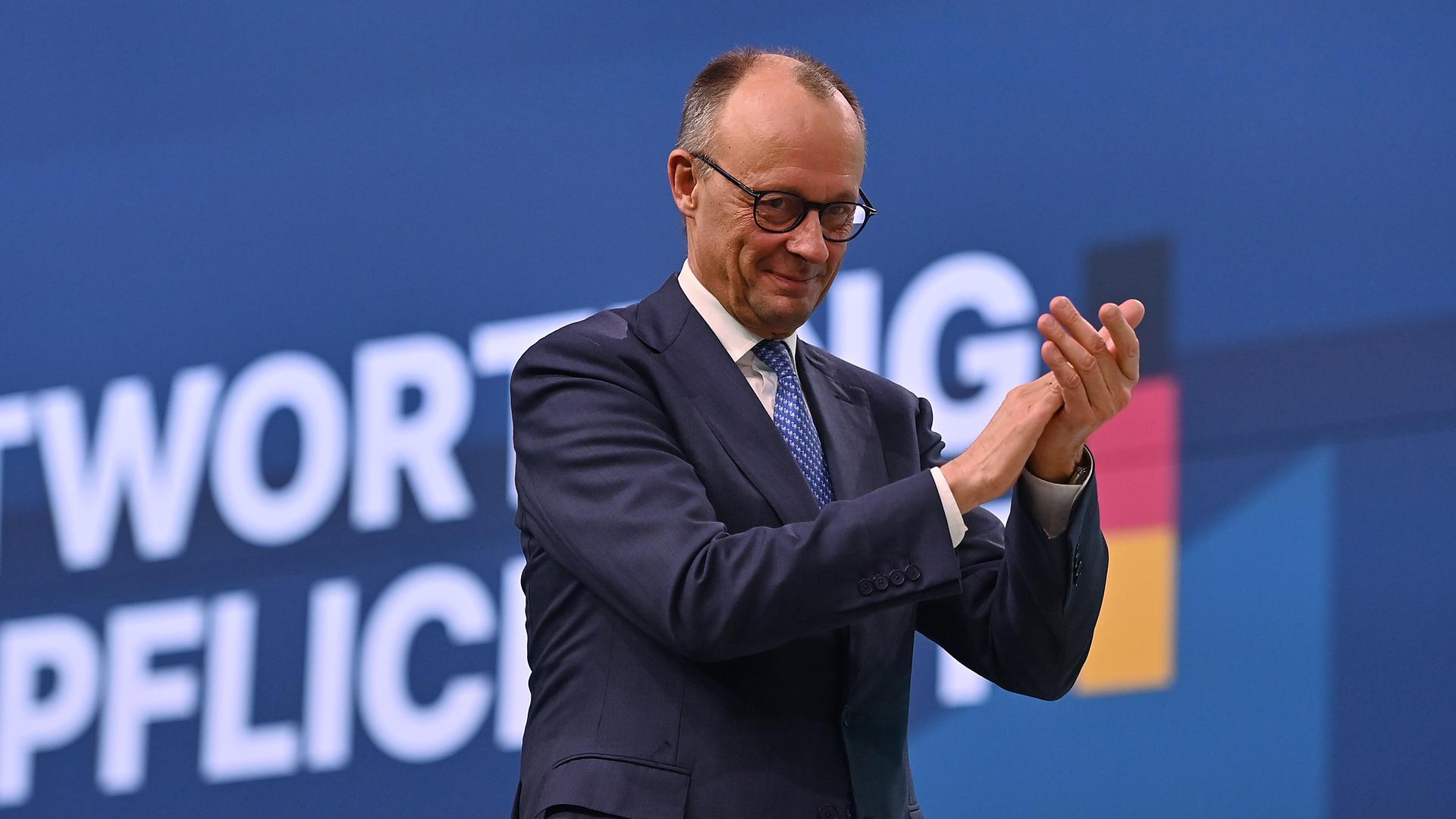 Bundes-kanzler Friedrich Merz steht vor einem blauen Hintergrund, lächelt und klatscht in die Hände. Er trägt einen Anzug.