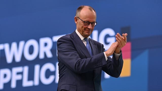 Bundes-kanzler Friedrich Merz steht vor einem blauen Hintergrund, lächelt und klatscht in die Hände. Er trägt einen Anzug.