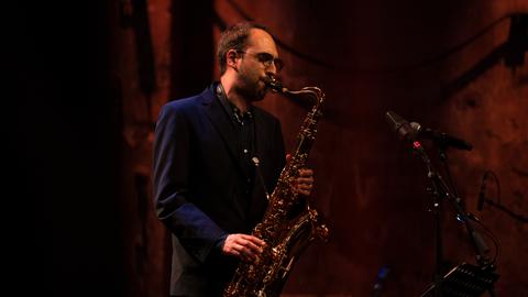 Ein junger Mann mit braunen Haaren und Brille spielt auf einem Saxofon.