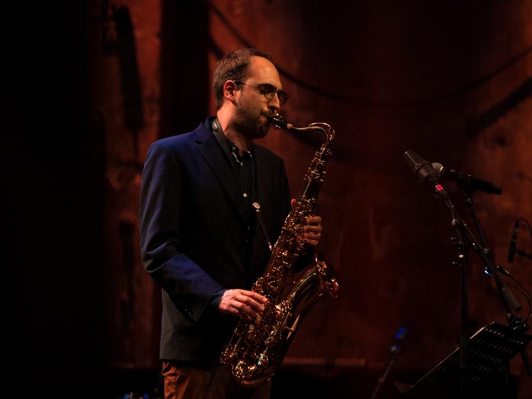 Ein junger Mann mit braunen Haaren und Brille spielt auf einem Saxofon.