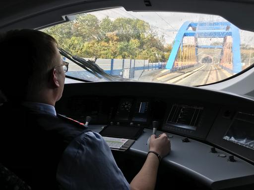Ein Lokführer sitzt im Führerstand und fährt einen ICE, vor der Scheibe ist ein blauer Brückenbogen zu sehen. 