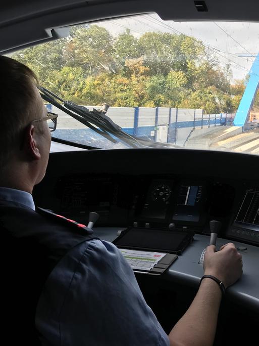Ein Lokführer sitzt im Führerstand und fährt einen ICE, vor der Scheibe ist ein blauer Brückenbogen zu sehen. 
