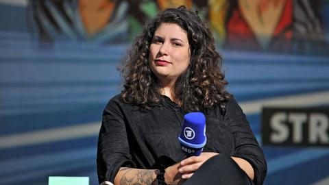 Jasmin Schreiber sitzt in schwarzer Kleidung und mit langen schwarzen Haaren auf einem Podium und hält ein Mikrofon in der Hand.