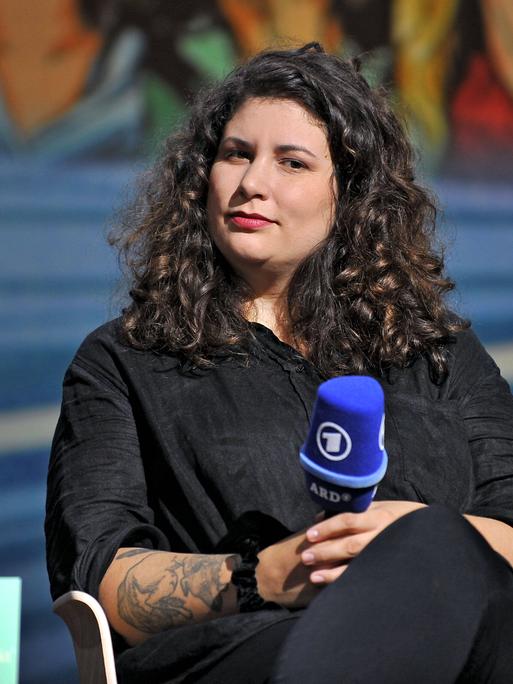 Jasmin Schreiber sitzt in schwarzer Kleidung und mit langen schwarzen Haaren auf einem Podium und hält ein Mikrofon in der Hand.