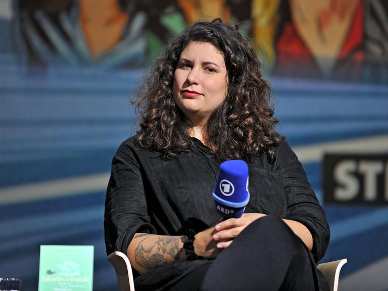 Jasmin Schreiber sitzt in schwarzer Kleidung und mit langen schwarzen Haaren auf einem Podium und hält ein Mikrofon in der Hand.