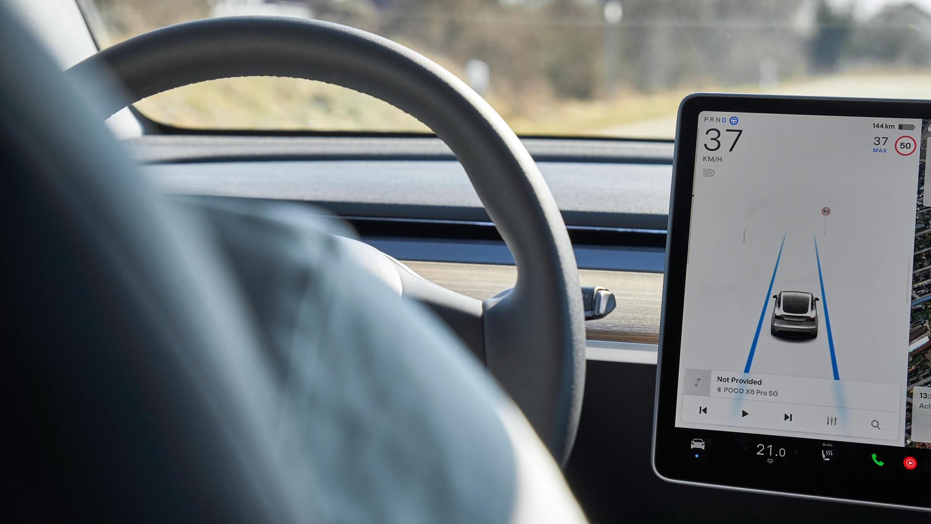 Das Display eines Tesla Model Y zeigt den Autopilot-Modus mit Spurführung. Das Fahrzeug fährt autonom auf einer Landstraße, während der Fahrer nicht eingreift. 
