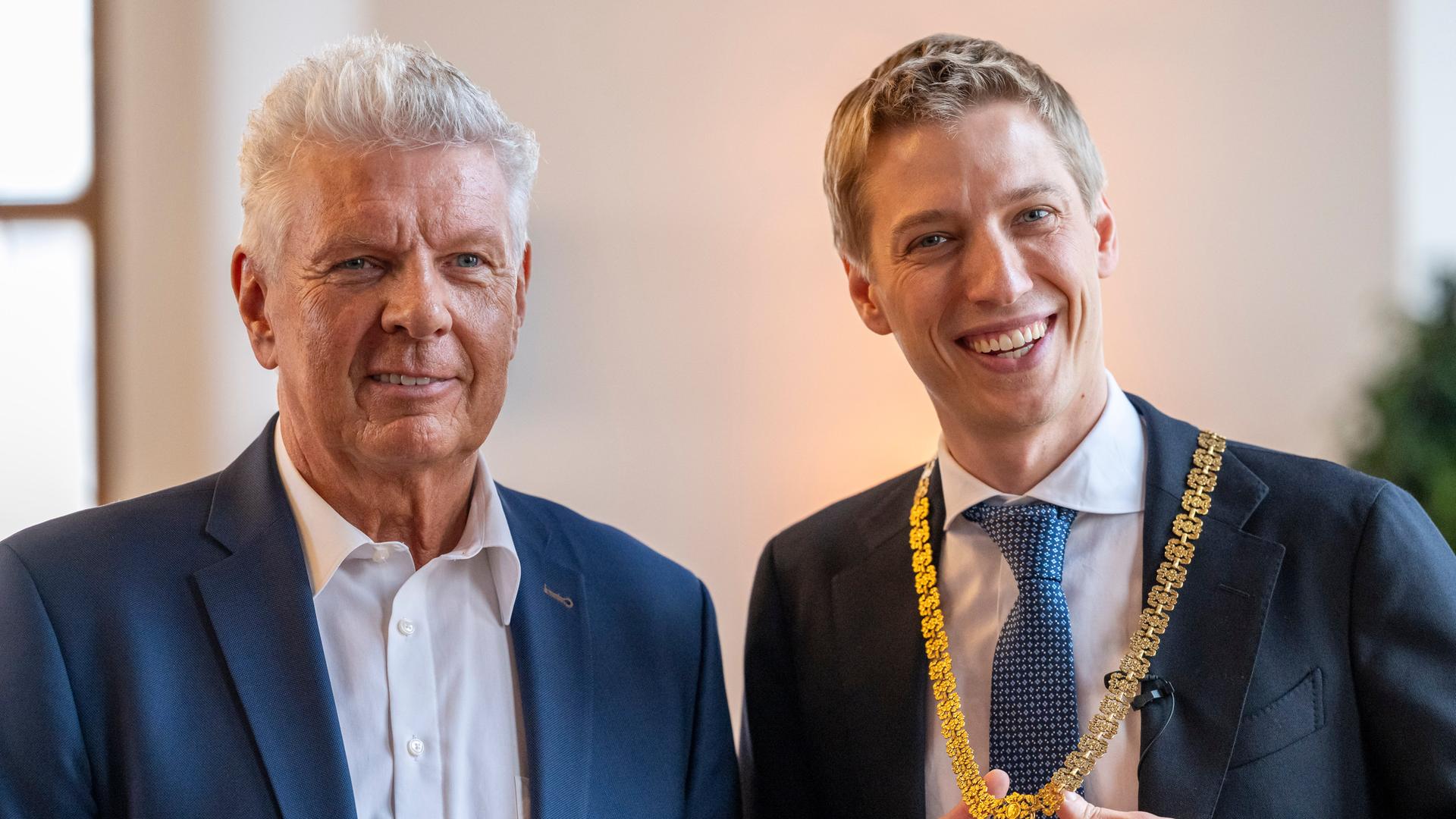 Der neue Oberbürgermeister Dominik Krause steht während seiner Amtseinführung im Großen Sitzungssaal des Rathauses neben seinem Amtsvorgänger Dieter Reiter (l). In der Hand hält er seine Amtskette. Er lacht.