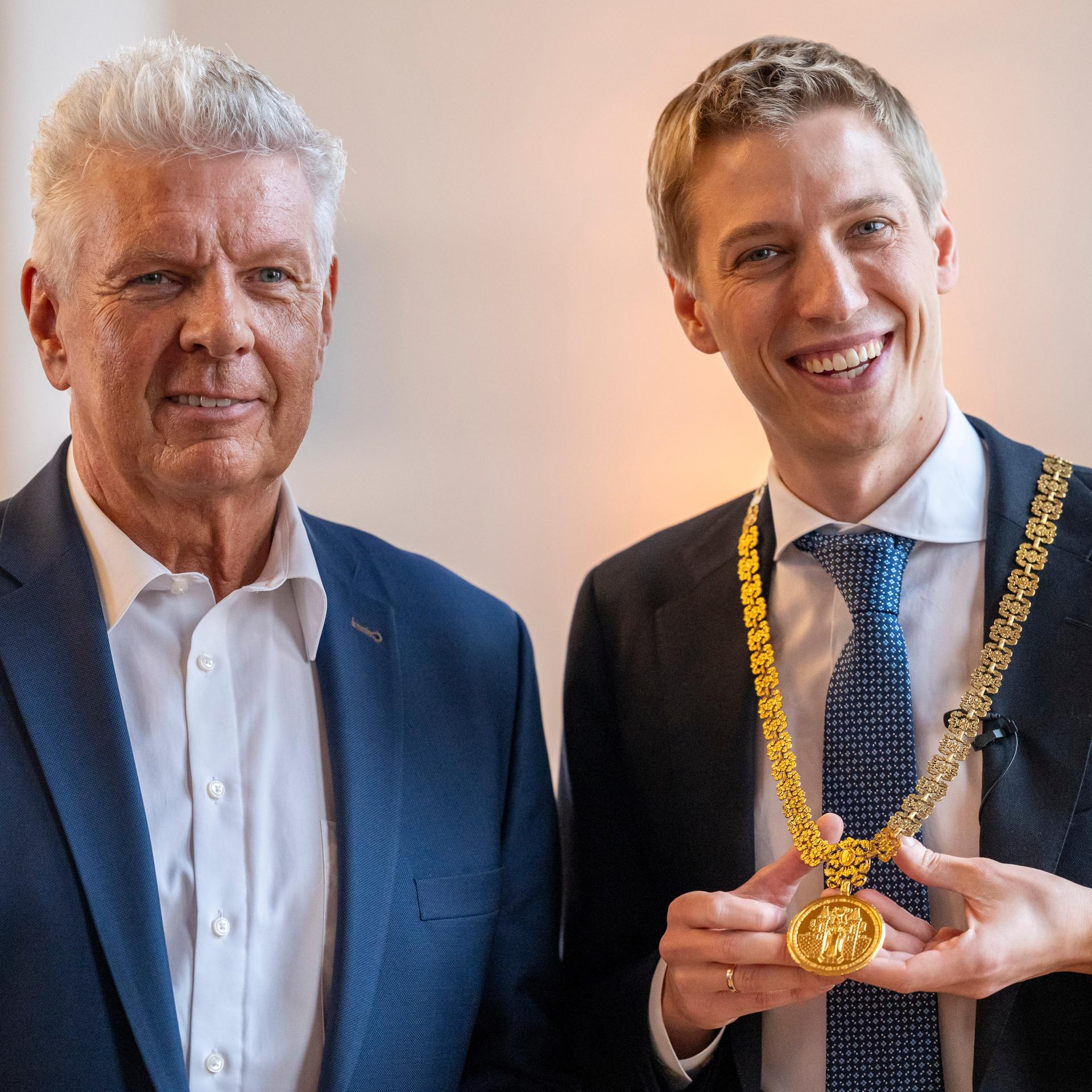Der neue Oberbürgermeister Dominik Krause (r, Grüne) steht während seiner Amtseinführung im Großen Sitzungssaal des Rathauses neben seinem Amtsvorgänger Dieter Reiter (l). In der Hand hält er seine Amtskette.