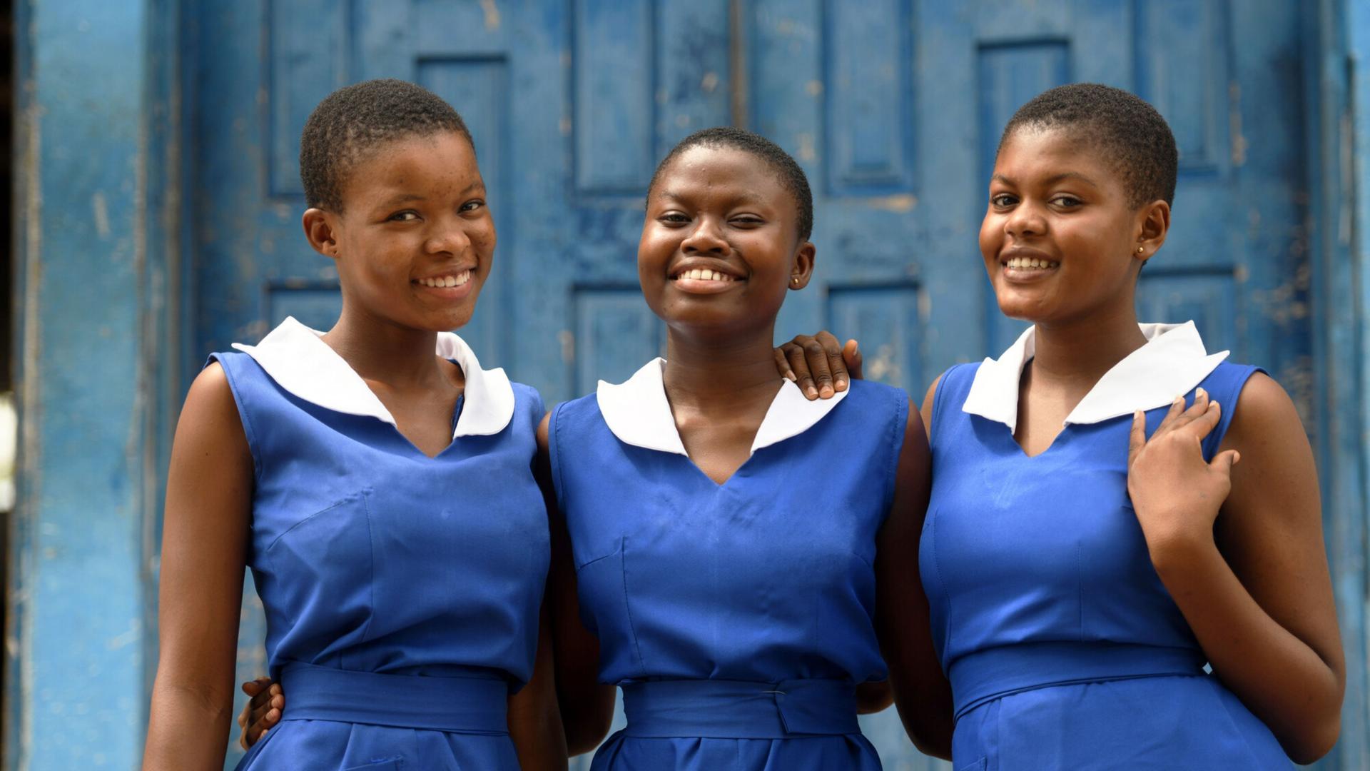 Drei Mädchen aus dem Land Ghana in Afrika. Die Mädchen haben die gleiche Kleidung an und lachen in die Kamera. Sie stehen in einer Reihe und haben ihre Arme umeinander gelegt