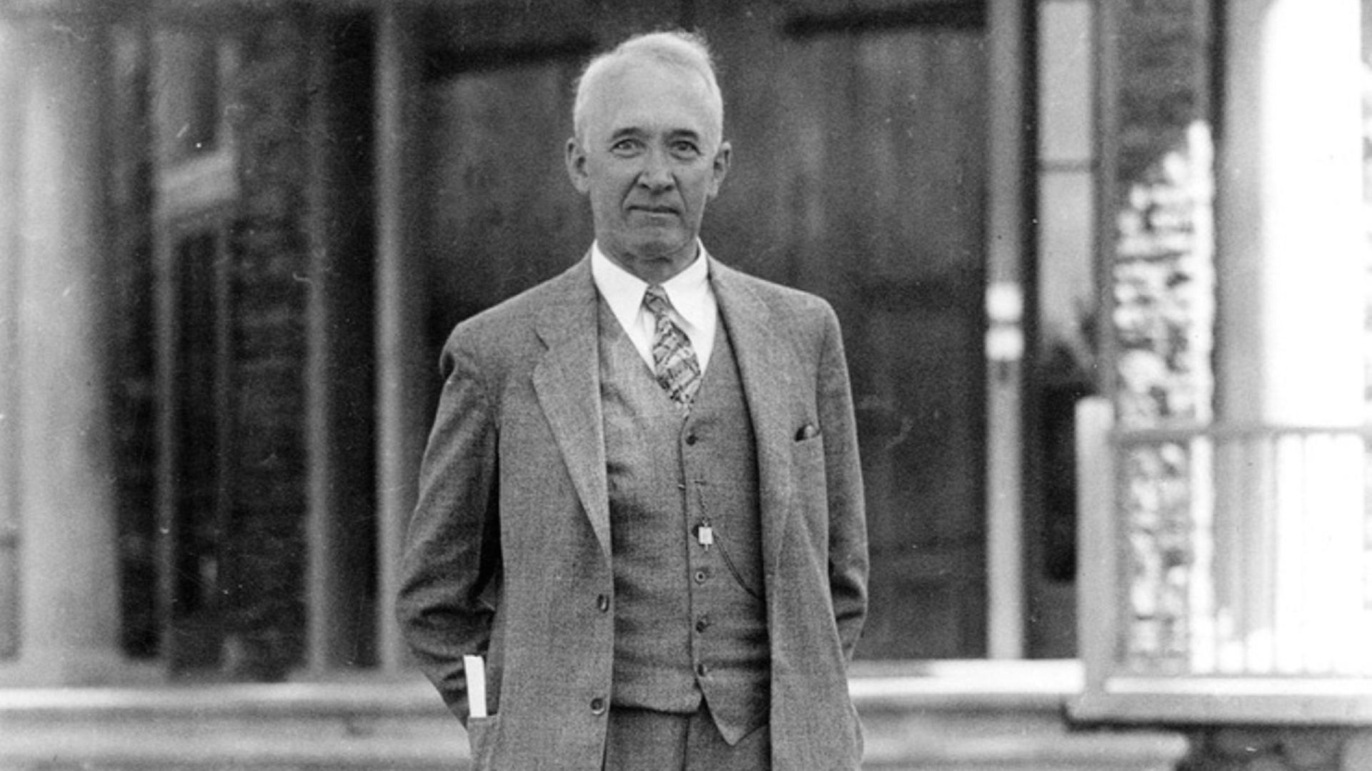 Ein Schwarz-Weiß-Bild zeigt den Astronomen Vesto Melvin Slipher, der in einem grauen Anzug mit Weste und Krawatte vor einem Gebäude für ein Foto posiert. Ein Schwarz-Weiß-Bild zeigt den Astronomen Vesto Melvin Slipher, der in einem grauen Anzug mit Weste und Krawatte vor einem Gebäude für ein Foto posiert.