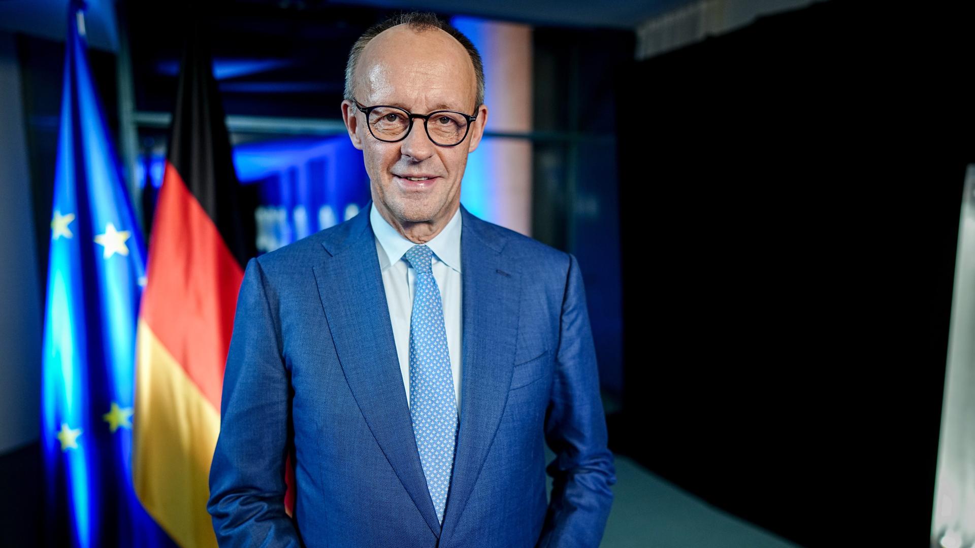 Bundeskanzler Friedrich Merz steht vor einer europäischen und einer deutschen Flagge und lächelt in die Kamera. 