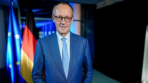Bundeskanzler Friedrich Merz steht vor einer europäischen und einer deutschen Flagge und lächelt in die Kamera. 