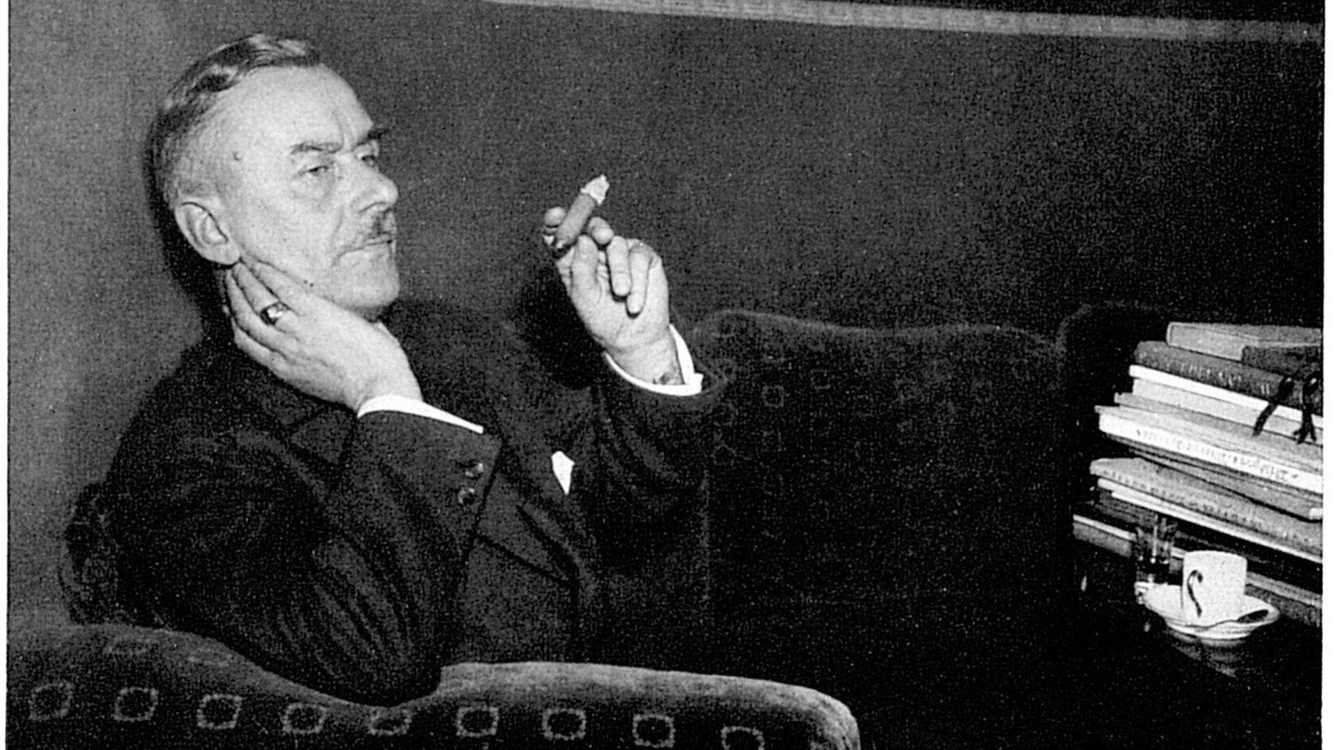Der deutsche Schriftsteller und Nobelpreisträger von 1929, Thomas Mann, sitzt auf einem Sofa mit einer Zigarre in der Hand, auf dem Tisch vor ihm Bücher und eine Tasse. 