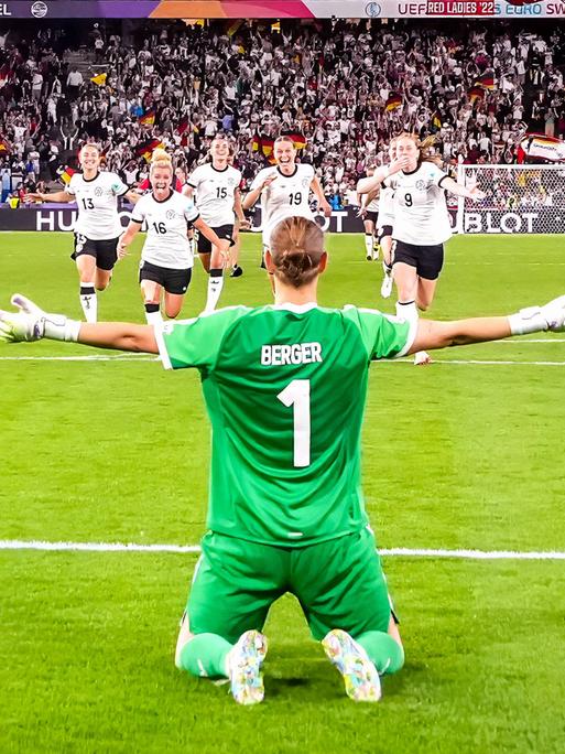 Rückansicht von Ann-Katrin Berger, die vor ihrem Tor kniet und die Arme ihrer Mannschaft entgegenstreckt, die auf sie zurennt.